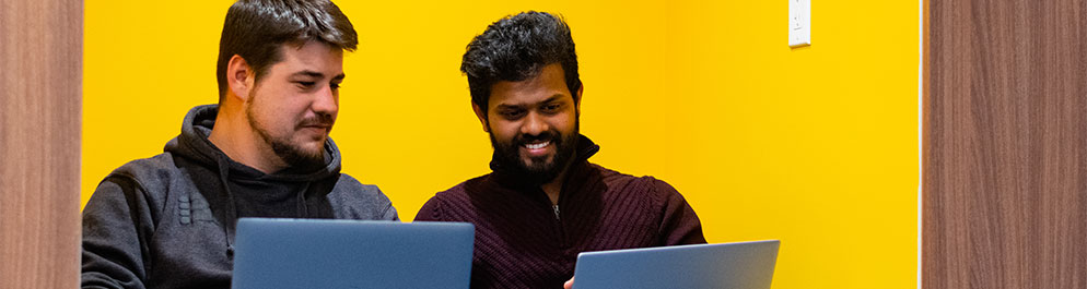 Two students sitting while using laptops and smiling