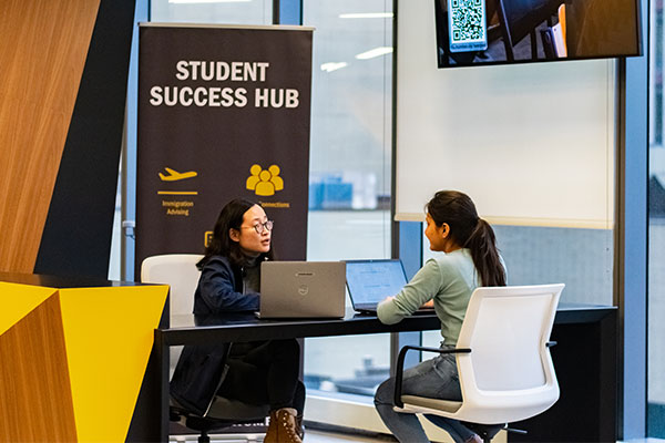 Two students sitting at a table talking in front of a student success hub sign